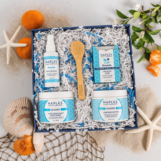 A curated beach-themed gift set from Naples Hand Cured Soap featuring four products in a navy blue box filled with white shredded paper. The set includes a Seaside body oil spray, a wooden spoon, a Spa Bliss Soap, a Body Butter and a Body Scrub.